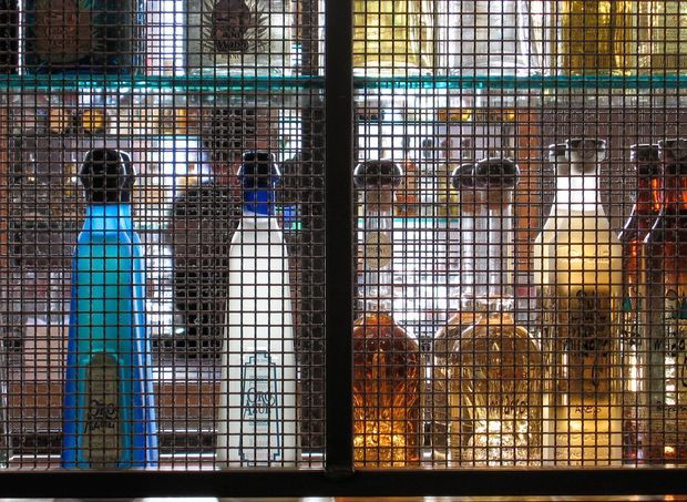 Close-up of a dark metal wire-mesh display cabinet filled with assorted bottles and containers, illuminated by warm and cool colored light. The image focuses on the gridded enclosure and its industrial retail or bar-like display contents.