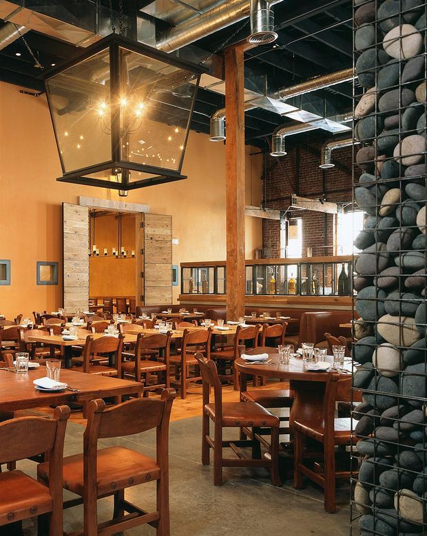 Interior view of a spacious industrial-style restaurant dining room with numerous solid timber tables and chairs, exposed ductwork and services, dark ceiling, timber structural posts, exposed brick walls, warm ochre plaster walls, and a large hanging lantern. A prominent column clad in rounded dark and light stones separates the dining area from the rear bar or service zone.