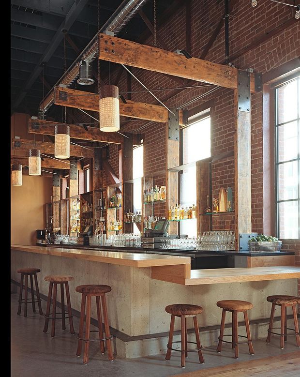 A spacious industrial-style bar interior with exposed red brick walls, heavy timber beams, steel columns, exposed ductwork, and tall black-framed windows. A long concrete and timber bar counter is lined with wooden stools, pendant lights, glassware, and illuminated liquor shelving. The scene has a warm, rustic character with strong architectural detailing and natural light.