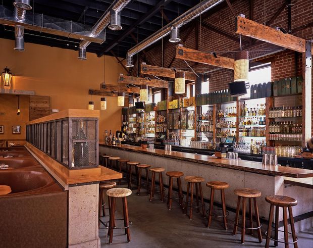 Interior view of a spacious industrial-style bar and restaurant. A long bar counter with numerous wooden stools runs along the right side, backed by extensive bottle display shelving and mirrors. Exposed red brick walls, dark exposed ceiling structure, galvanized metal ductwork, weathered timber beams, pendant lights, and concrete-look surfaces create a warm, rustic industrial atmosphere. Booth seating and a raised timber-and-metal partition occupy the foreground.