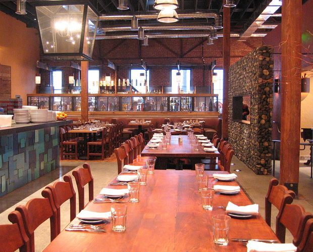 A spacious industrial-style restaurant interior with long communal wooden dining tables set with white plates, folded napkins and drinking glasses. Exposed red brick walls, dark exposed ceiling services, ductwork, timber posts, metal-framed glazing and pendant lighting define the space. A stone-clad feature wall and tiled bar counter add texture, while additional seating and a bar area are visible in the background.