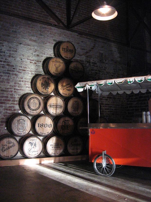 A dimly lit industrial-style interior with exposed dark brick walls and exposed timber roof trusses. A decorative stack of tequila barrels forms a feature display beside a bright red wheeled food or drinks cart with a striped canopy. A large metal pendant light hangs overhead, and strong sunlight falls across the dark concrete floor.