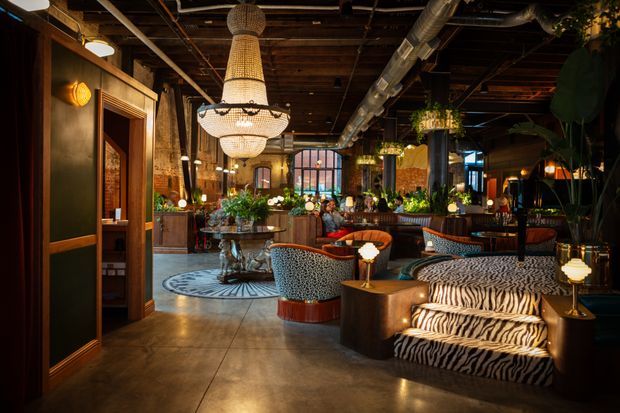 A richly layered, moody restaurant or hospitality interior with exposed dark timber ceiling structure, visible industrial ductwork, polished concrete flooring, dark wood and black wall finishes, and abundant tropical planting. The space features ornate woven pendant chandeliers, patterned upholstered lounge seating, zebra-print sofas, dining tables, a bar or service area, and warm atmospheric lighting. The overall design blends industrial architecture with eclectic, luxurious, vintage-inspired furnishings.