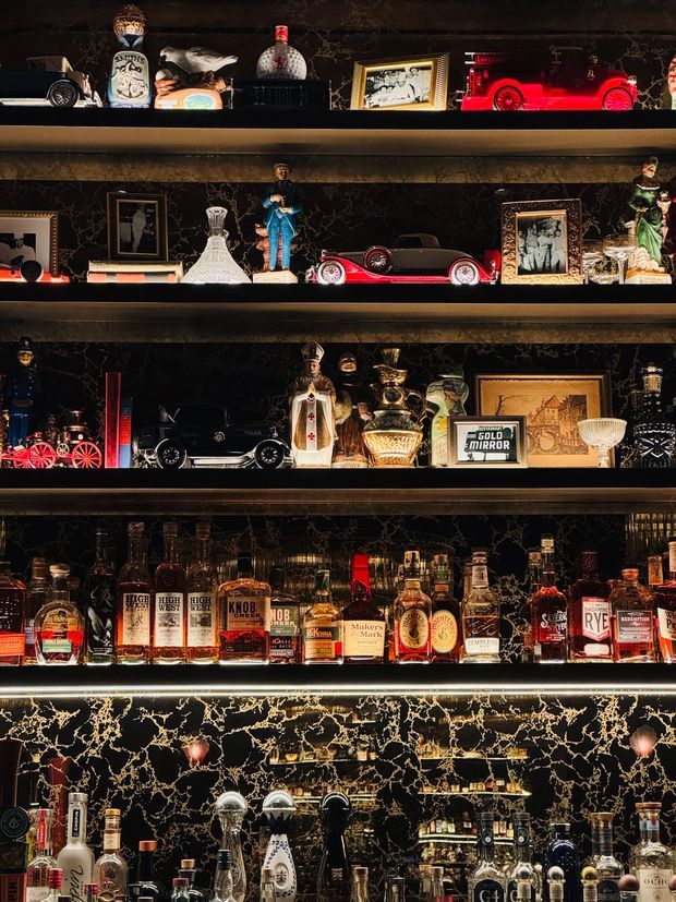 A dramatic, dark display wall with multiple illuminated shelves filled with liquor bottles, vintage toy cars, framed artwork, figurines, decorative glassware, and bar memorabilia. The lower section includes a bar counter or backbar area, while black marble-look surfaces with bold gold veining create a luxurious, moody setting.