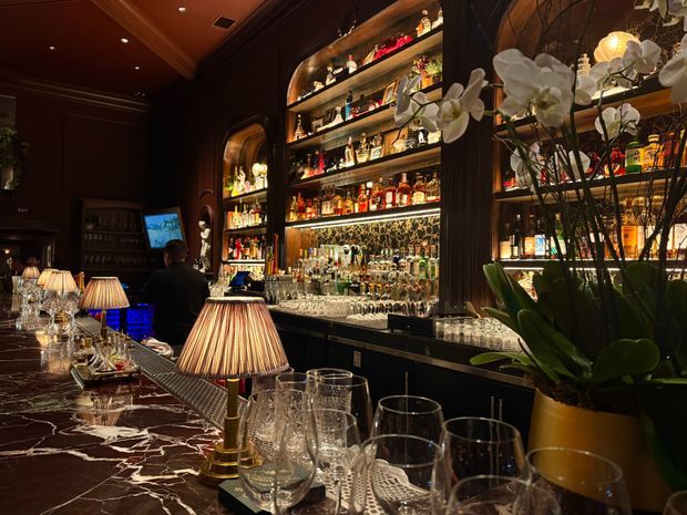 A dark, richly detailed cocktail bar interior with a long polished stone bar counter, illuminated back-bar shelving filled with liquor bottles and glassware, decorative table lamps, dark timber panelling, and orchids in the foreground. The atmosphere is intimate and luxurious, with a bartender seated or working behind the counter.