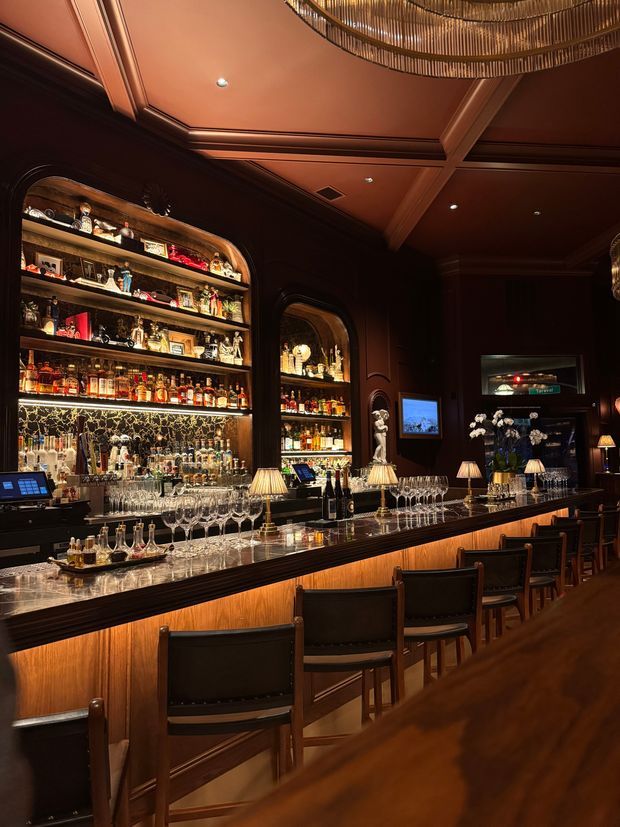 A richly detailed, dimly lit bar interior with a long marble-topped counter, dark timber cabinetry and wall panelling, illuminated liquor display shelving, leather bar stools, glassware, table lamps, and a coffered timber ceiling. The space has an elegant, intimate hospitality setting with warm amber lighting and reflective metallic and glass details.