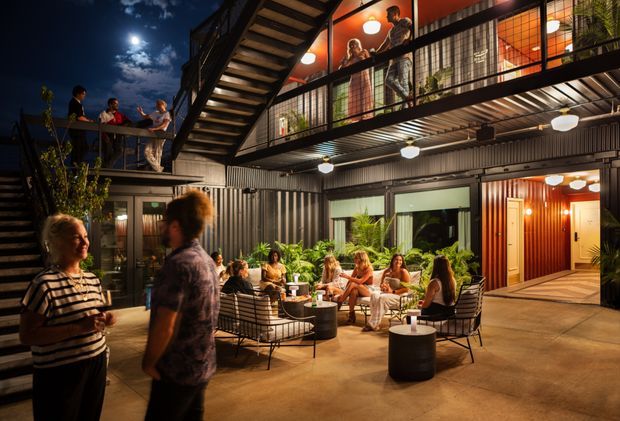 Night-time view of a lively multi-level courtyard or hospitality-style communal space, with people socialising around outdoor lounge seating and tables. The building features dark corrugated metal walls, extensive black-framed glazing, exposed steel balcony railings, an external stair, warm pendant and wall lighting, and planted greenery. Upper-level balconies overlook the central courtyard.