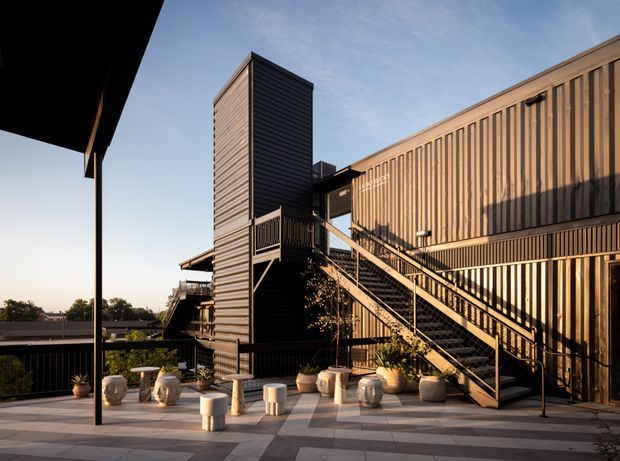 Exterior rooftop terrace of a contemporary multi-level building, featuring dark vertical and horizontal cladding, an exposed external staircase and balcony, and a paved outdoor seating area with planters and stools. Warm late-day sunlight creates strong shadows across the facade and terrace.