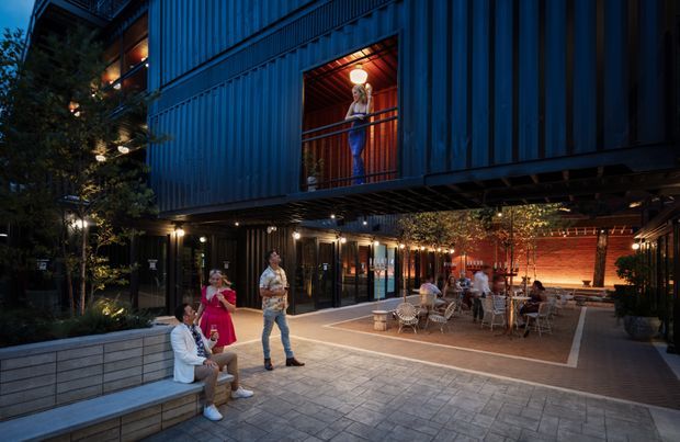 A dusk architectural view of a contemporary multi-level commercial courtyard or hospitality space formed by blue corrugated-metal-clad upper volumes around a paved outdoor dining area. Warm lighting illuminates the brick feature wall, glazed ground-floor frontage, balconies, landscaping, and outdoor café seating, with several people occupying the courtyard.