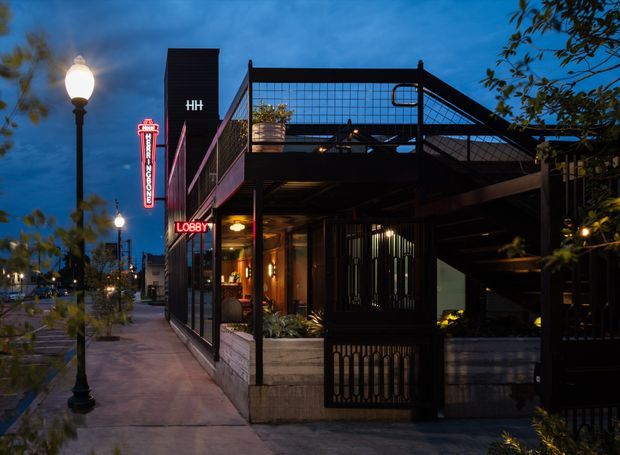 A dusk exterior view of a contemporary multi-level commercial building on a city sidewalk. The dark facade incorporates extensive glazing, black metal framing, an elevated terrace or balcony with mesh guardrails, illuminated signage, warm interior lighting, planted window boxes, and street lamps along the footpath.