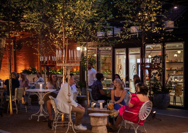 A warmly lit outdoor dining and social area at night, with patrons seated at small café tables and ornate chairs beneath mature trees. The space fronts a glazed commercial venue with illuminated shop windows and signage, while brick walls, planters, and atmospheric lighting create an inviting urban courtyard or streetside dining setting.