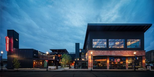 Evening exterior view of a contemporary commercial building or hospitality venue with a dramatic projecting flat roof, dark metal cladding, exposed brick lower facade, large multi-pane industrial-style windows, and warm interior lighting. Adjacent low-rise commercial structures and illuminated signage line the streetscape.