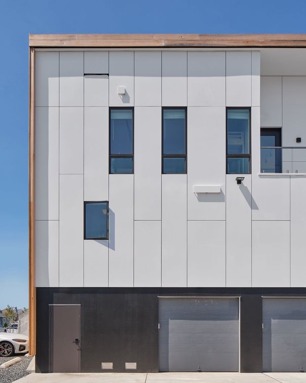 A close-up exterior view of a contemporary multi-storey building facade under a clear blue sky. The upper facade is clad in pale grey rectangular panels with narrow vertical windows, small projecting elements, and a balcony opening. A dark lower level contains multiple garage doors and a pedestrian entry door, while timber-toned trim frames the roofline and side edge.