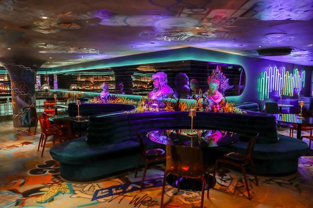 A dark, highly stylised cocktail bar or lounge with curved upholstered banquette seating around low tables, a long illuminated bar, mirrored and glossy surfaces, colourful sculptural decor, neon lighting, and vivid projected graphic artwork across the ceiling and floor. The space has a theatrical, immersive nightlife atmosphere with purple, blue, pink, and turquoise lighting.