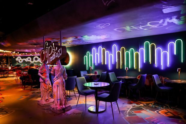 A dark, vividly lit themed hospitality interior resembling a cocktail bar or lounge. Small round tables and upholstered dining chairs are arranged across a patterned floor, with a long wall featuring multicoloured neon tube lighting and projected or painted graphic artwork across the ceiling and floor. Sculptural decorative figures and a hanging sign contribute to the theatrical, retro-pop atmosphere.