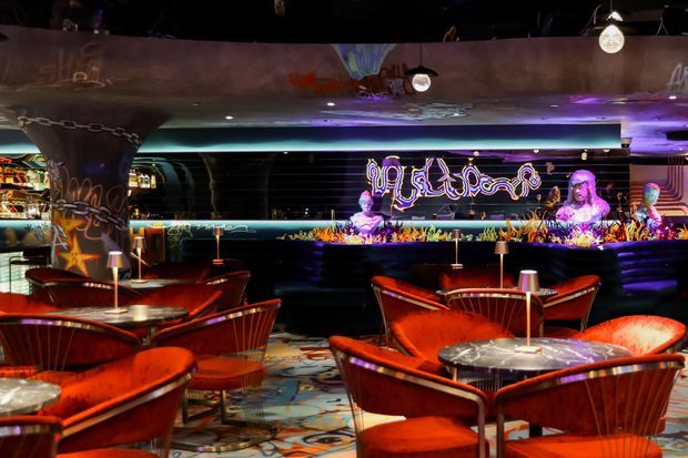 A dimly lit retro-inspired commercial lounge or bar with vivid red upholstered booth seating, small round tables, metallic accents, colourful patterned flooring, and a long illuminated bar counter. Graffiti-style artwork, neon signage, purple-blue ambient lighting, and decorative pendant lights create a dramatic nightlife atmosphere.
