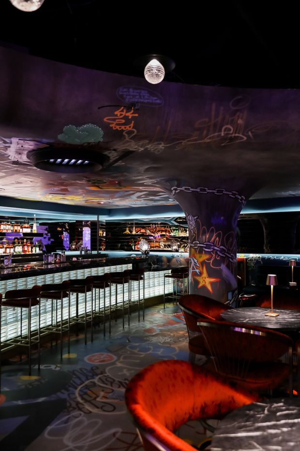 A dramatic, dimly lit commercial bar and lounge interior with a long illuminated bar, rows of high stools, reflective surfaces, and low lounge seating. Graffiti-style projections and murals cover the ceiling, walls, central column, and floor, while red-orange upholstered chairs, small table lamps, and dark finishes create an energetic nightclub atmosphere.