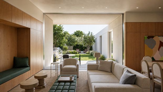 A refined contemporary living room with warm timber wall panelling and built-in green bench seating, a large neutral sectional sofa, sculptural coffee tables, and an open connection to a landscaped courtyard. A dining area with curved-back chairs and colourful abstract artwork is visible to the right. The room has a low, smooth ceiling with recessed lighting and broad floor-to-ceiling openings that bring in abundant daylight.