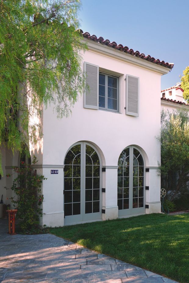 Exterior view of a refined two-storey stucco building with pale pink rendered walls, dark red curved roof tiles, grey louvred shutters, and tall arched multi-pane glazed doors. A paved walkway, lawn, mature planting, and a small address plaque frame the facade.