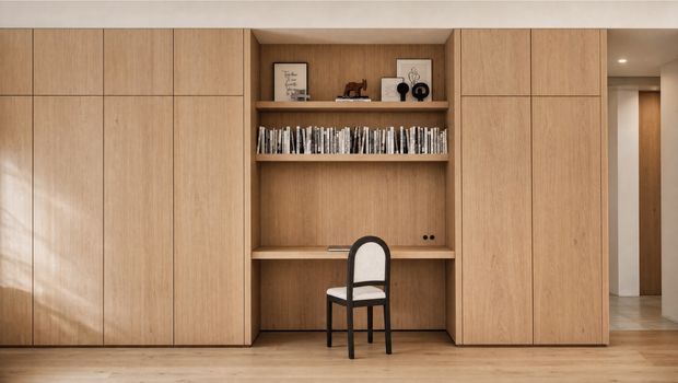 A warm, minimalist built-in timber wall unit with full-height cabinetry framing an open shelving and compact desk niche. The central recess contains books, framed decor and a small black chair, while pale timber flooring and a simple white ceiling create a calm, refined interior.