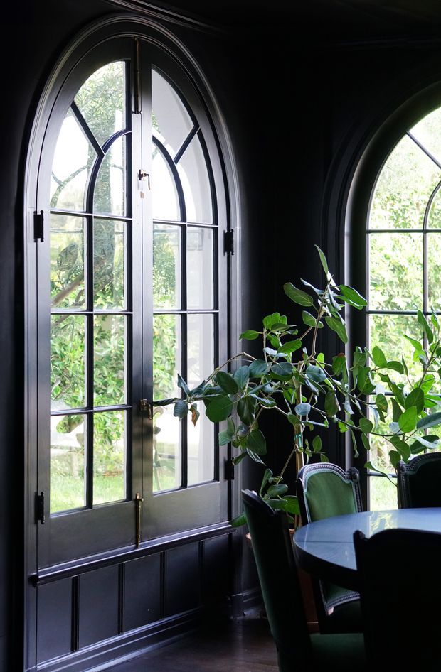 A dark, elegant dining area with dramatic black wall panelling and matching arched French-style windows and doors. Daylight filters through the divided glazing, highlighting lush greenery outside and a leafy indoor plant. A round dining table with upholstered green chairs occupies the foreground. The room has ornate arched joinery and a moody, refined atmosphere.