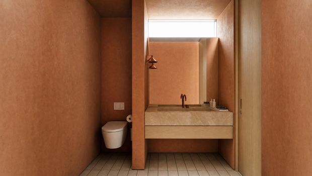 Compact, warm-toned bathroom with terracotta plaster walls, a wall-mounted toilet, and a built-in vanity with a stone benchtop, integrated basin and tapware. A large mirror and concealed shelving are set above the vanity, with timber cabinetry and a tiled floor.