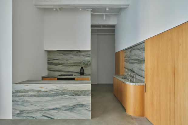 A compact contemporary kitchen with dramatic veined stone used across the island, benchtops, splashbacks and the feature wall behind the cooktop. Warm timber cabinetry contrasts with the cool stone, while exposed white ceiling services and a narrow doorway add an understated commercial or loft-like character.
