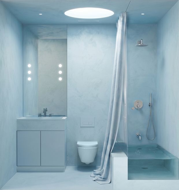 A compact monochromatic pale-blue bathroom with seamless textured wall and floor finishes. It includes a floating double-door vanity with integrated basin and wall-mounted taps, a mirrored cabinet, wall-hung toilet, walk-in shower with a glass partition and white curtain, handheld shower, rain showerhead, recessed lighting, and a large circular ceiling light.