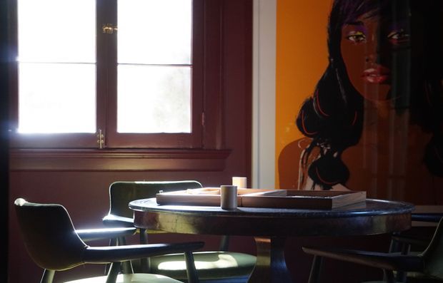 A moody dining area with a round dark wood table, several mid-century-style upholstered dining chairs, and a large colorful portrait artwork on the wall. Strong daylight enters through a dark timber-framed window, creating high contrast against warm orange and brown tones.