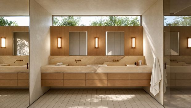 A spacious contemporary bathroom with a long double vanity in warm timber cabinetry, a pale stone benchtop and integrated basin, and brushed metal tapware. The vanity wall features vertical timber panelling, illuminated mirrors, and slim wall sconces. Large windows bring in abundant natural light, while full-height mirrors reflect the room and a towel hangs beside the vanity. The floor is finished in small light beige tiles.