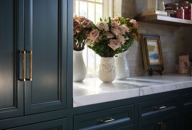 Close-up of a refined built-in dark blue-green cabinetry installation with raised-panel doors and brass hardware. A white marble countertop sits beneath a window, styled with ceramic vases of blush flowers, framed artwork, and shelving with jars. The space has a classic, elegant, softly lit character.