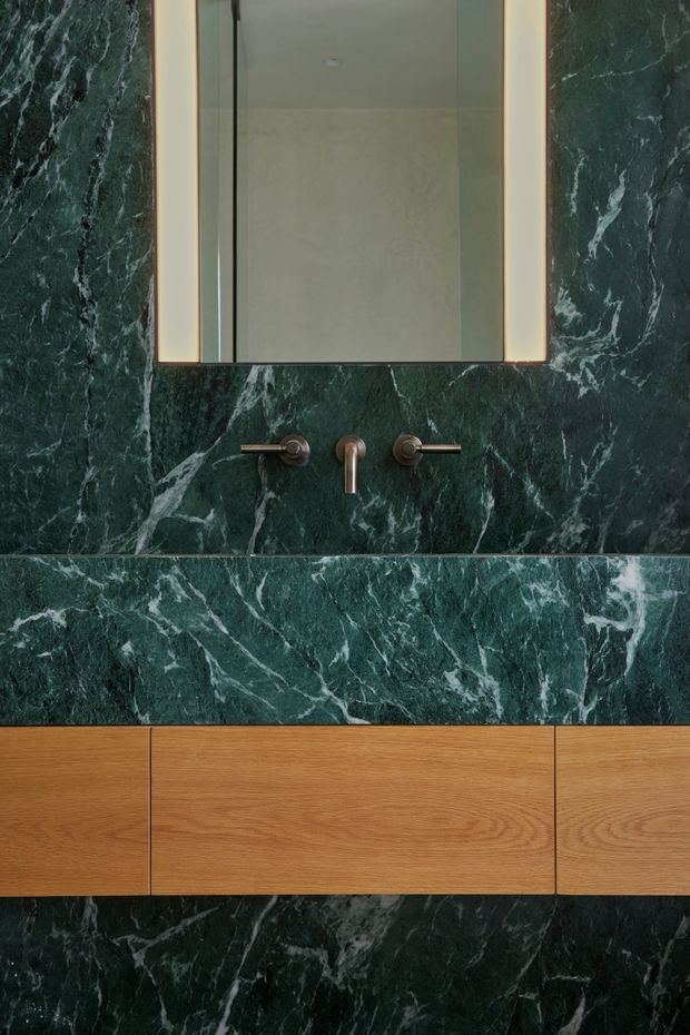 Close-up view of a refined bathroom vanity featuring dramatic dark green veined stone across the wall and benchtop, a floating warm timber vanity with integrated drawers, wall-mounted brushed metal tapware, and a rectangular illuminated mirror.