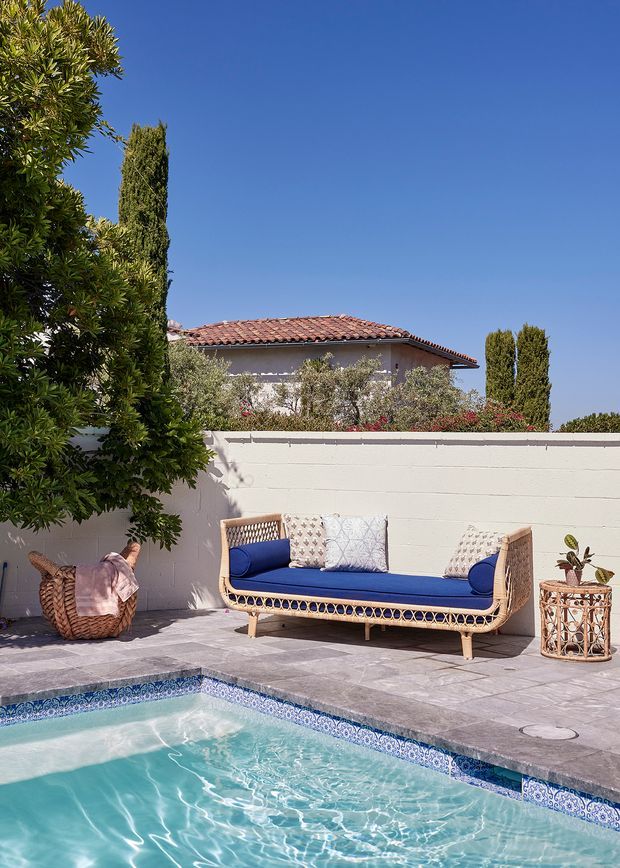 Sunny outdoor pool area with turquoise water, patterned blue-and-white pool tile, and a paved terrace furnished with a rattan sofa upholstered in deep blue and patterned cushions. A white rendered masonry wall, mature greenery, tall cypress trees, and a terracotta-tiled roof create a Mediterranean-inspired residential setting.