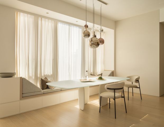 A bright, refined dining area with a built-in window seat beneath full-height sheer curtains, a sculptural pale dining table, upholstered dining chairs, and a cluster of metallic globe pendant lights. The room has warm pale timber flooring, off-white walls and fixed cabinetry, with a minimalist, softly elegant interior.
