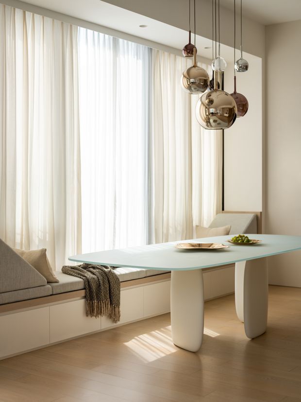 A refined, light-filled dining area with a pale glass-topped table supported by sculptural rounded legs, positioned beside a built-in window seat with upholstered cushions. Floor-to-ceiling sheer curtains diffuse sunlight across the pale timber floor. A cluster of reflective spherical pendant lights hangs above the table, creating a soft contemporary and luxurious atmosphere.
