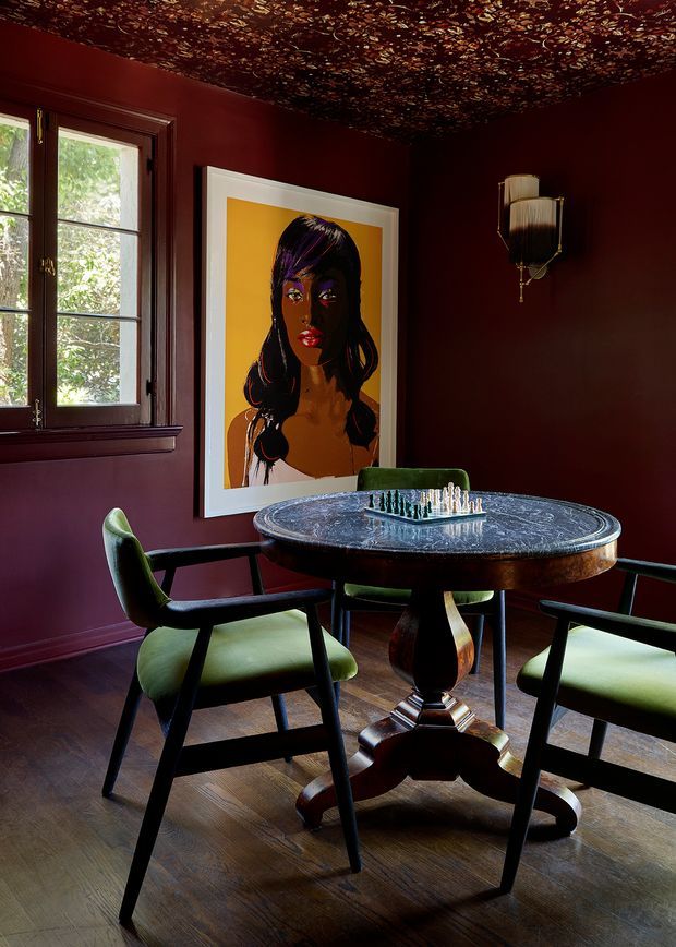 A richly coloured, moody dining room with deep burgundy walls and a dark patterned decorative ceiling. A round pedestal dining table with a dark stone-like top is surrounded by green upholstered mid-century-style chairs, with a chess set as the centrepiece. Tall timber-framed windows bring in daylight, while a large colourful portrait, sculptural wall sconce, and dark timber flooring add character.