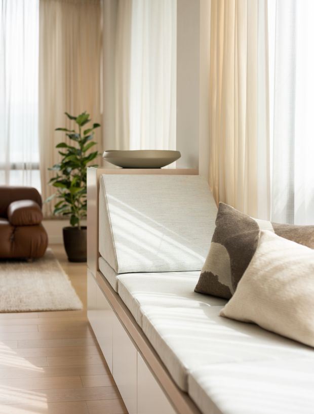 Bright, serene living room with a built-in white upholstered window seat or daybed, neutral cushions, a shallow ceramic bowl, sheer floor-to-ceiling curtains, pale timber flooring, and a potted indoor tree. A brown leather lounge chair and soft rug are visible in the background. The room has abundant filtered natural light and a refined neutral palette.