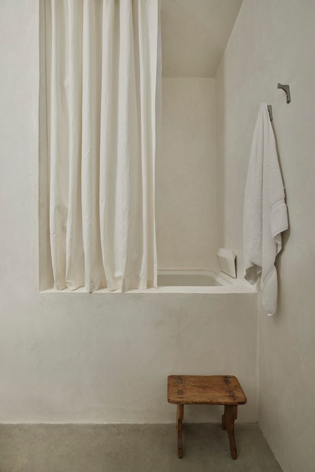 A compact, softly lit bathroom with a built-in bathtub or shower-bath enclosure behind a white fabric curtain. The space has pale plastered walls, a muted grey concrete-like floor, a white towel on a wall hook, and a small rustic wooden stool. The restrained palette and simple fixtures create a quiet, minimalist atmosphere.
