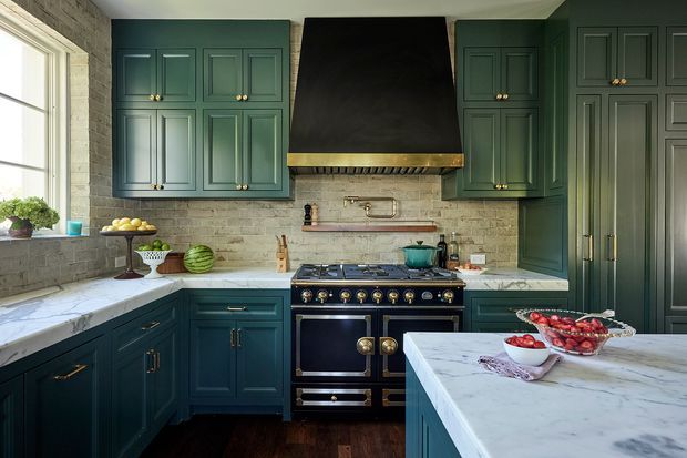 A richly detailed kitchen with deep teal-green raised-panel cabinetry, brass hardware, a large black range cooker and hood, pale textured brick backsplash, white marble-look benchtops, and dark timber flooring. Natural light enters from a large window, creating a refined yet warm and traditional atmosphere.