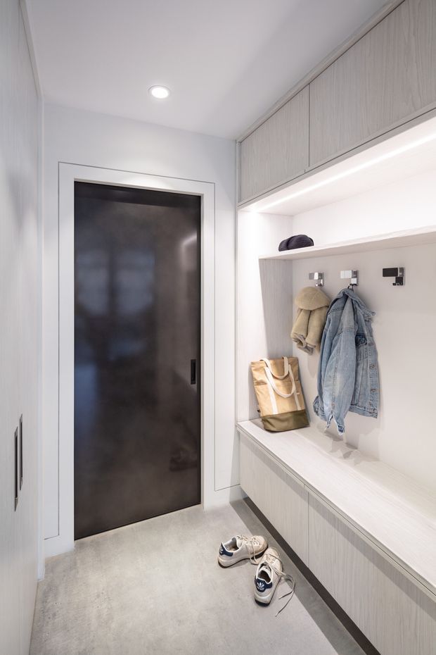 A bright, narrow entryway or mudroom with built-in light wood-grain cabinetry, an integrated bench, open shelf, coat hooks, and concealed overhead storage. A dark flush door contrasts with the pale cabinetry and smooth grey floor. Recessed ceiling lighting and under-shelf illumination create a clean, highly functional contemporary interior.