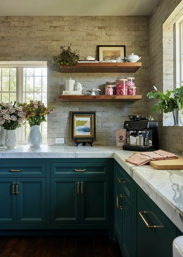 A compact, richly detailed kitchen corner with deep teal-green shaker cabinetry, white marble-look countertops, exposed light brick walls, dark timber flooring, and warm brass hardware. Open timber shelves display ceramics, framed artwork, jars, and plants. A large window brings in natural light, while a coffee machine and prep area sit on the right-hand counter.