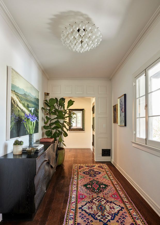 A bright, refined residential hallway with dark timber flooring, white paneled walls and ornate crown moulding. A long patterned runner leads past a black console with plants and artwork toward an open doorway. A crystal chandelier, large divided-light window, framed art and a tall indoor plant add decorative character.