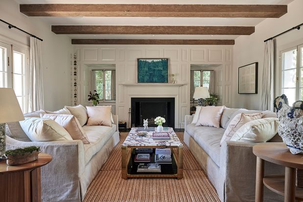 A bright, refined living room with two facing upholstered sofas arranged around a central wood-and-glass coffee table and a fireplace. The room features white paneled walls, exposed dark timber ceiling beams, tall curtained windows, a neutral woven rug, and soft blush and cream furnishings.