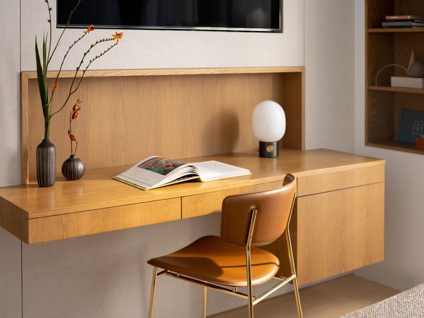 A compact, warmly lit built-in home office or study nook beneath a wall-mounted television. The room features a floating timber desk with integrated shelving and cabinetry, a timber back panel, an open book, decorative vases, and a brown leather chair with a slender brass-toned frame. Additional timber display shelving is visible at right, with a neutral wall and partial textured rug completing the scene.