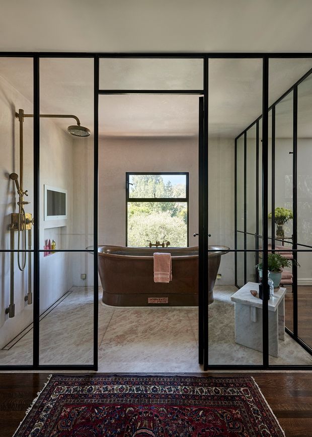 A refined bathroom viewed through black metal-framed glass partitions and a hinged glass door. The room features a freestanding copper-toned bathtub centered beneath a window, an exposed shower with brass fittings on the left, a pale stone or marble-look floor, and a marble vanity with a vessel basin on the right. Dark timber flooring and a richly patterned Persian-style rug sit outside the bathroom.