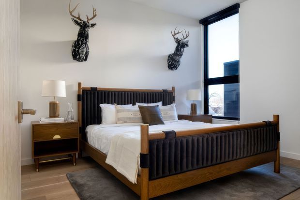 A refined bedroom with a dark wood four-poster-style bed featuring vertically channelled upholstered headboard and footboard panels. White bedding, neutral walls, timber flooring, a dark area rug, matching bedside tables and lamps create a balanced contemporary interior. Black decorative deer-head wall sculptures and a large black-framed window add graphic contrast.