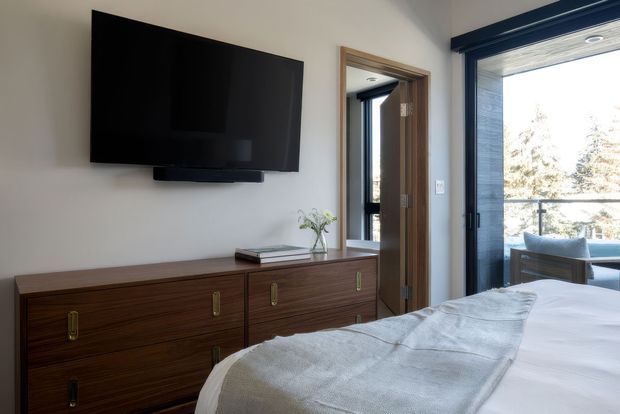 A bright, contemporary bedroom with a large wall-mounted television above a dark timber dresser, a neatly made bed in the foreground, and a glazed doorway opening to a balcony or exterior view. The room has white walls, timber door and trim, and abundant natural light.