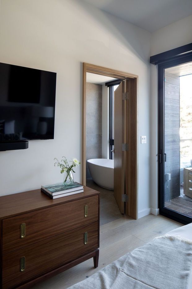 A refined bedroom interior viewed toward an open ensuite bathroom. The room features a mid-century-style dark timber dresser with brass pulls, a wall-mounted black television, pale timber flooring, white walls, and a large black-framed glazed door. Through the timber doorway, a freestanding white bathtub is visible in the bathroom. Natural light creates a calm, sophisticated atmosphere.