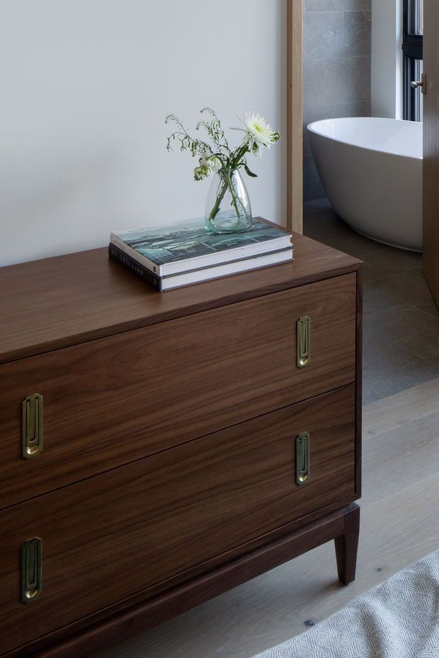 A refined bedroom or dressing area featuring a dark mid-century-style timber chest of drawers with elongated brass handles. A stack of books and a glass vase with flowers sit on top. Through the open doorway, a freestanding white bathtub is visible in a tiled bathroom, alongside dark-framed glazing. The space has warm timber joinery, pale walls, and a light timber floor.