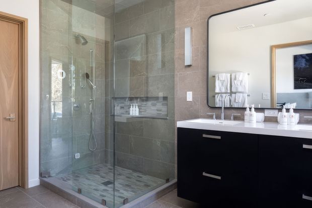 Contemporary bathroom with a frameless glass walk-in shower, grey large-format wall tiles, mosaic shower floor, and a floating black vanity with a white benchtop and double basins. A large rounded-corner mirror, wall-mounted lighting, timber door, and neatly arranged towels and toiletries complete the refined, hotel-like space.